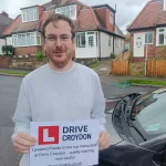 Learner holding a practical driving test pass certificate beside a Drive Croydon car
